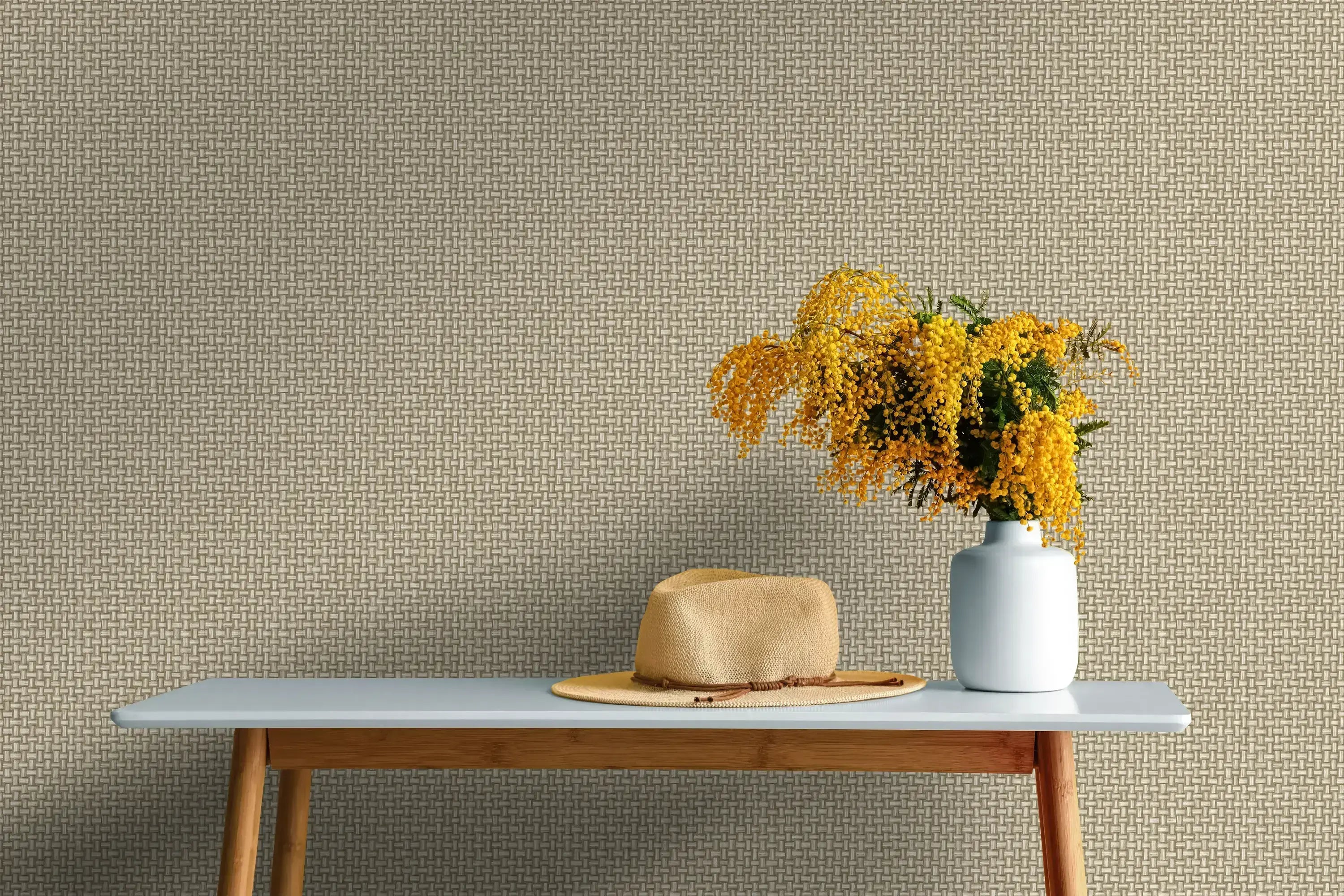 Biderbost Weave - Beige tapet som baggrund i et rum med en hvid vase med gule blomster og en stråhat på en træbordplade.