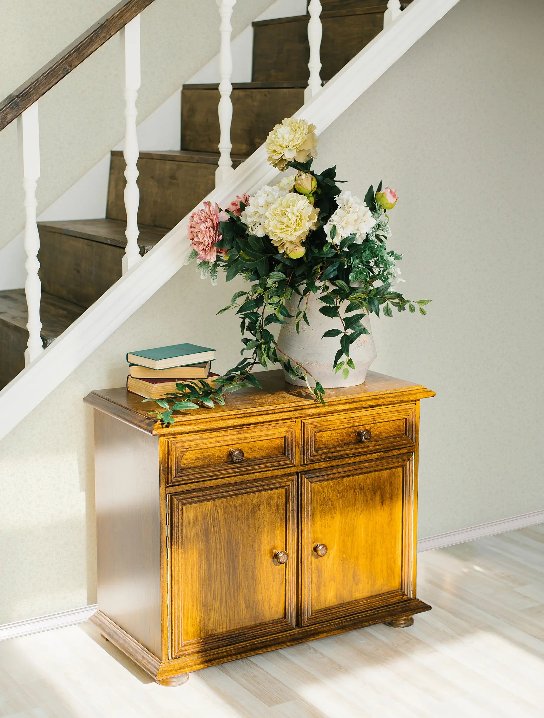 Catherine Floral - Ivory tapet i en indretning med en trækommode og blomster, der fremhæver det elegante blomstermønster.