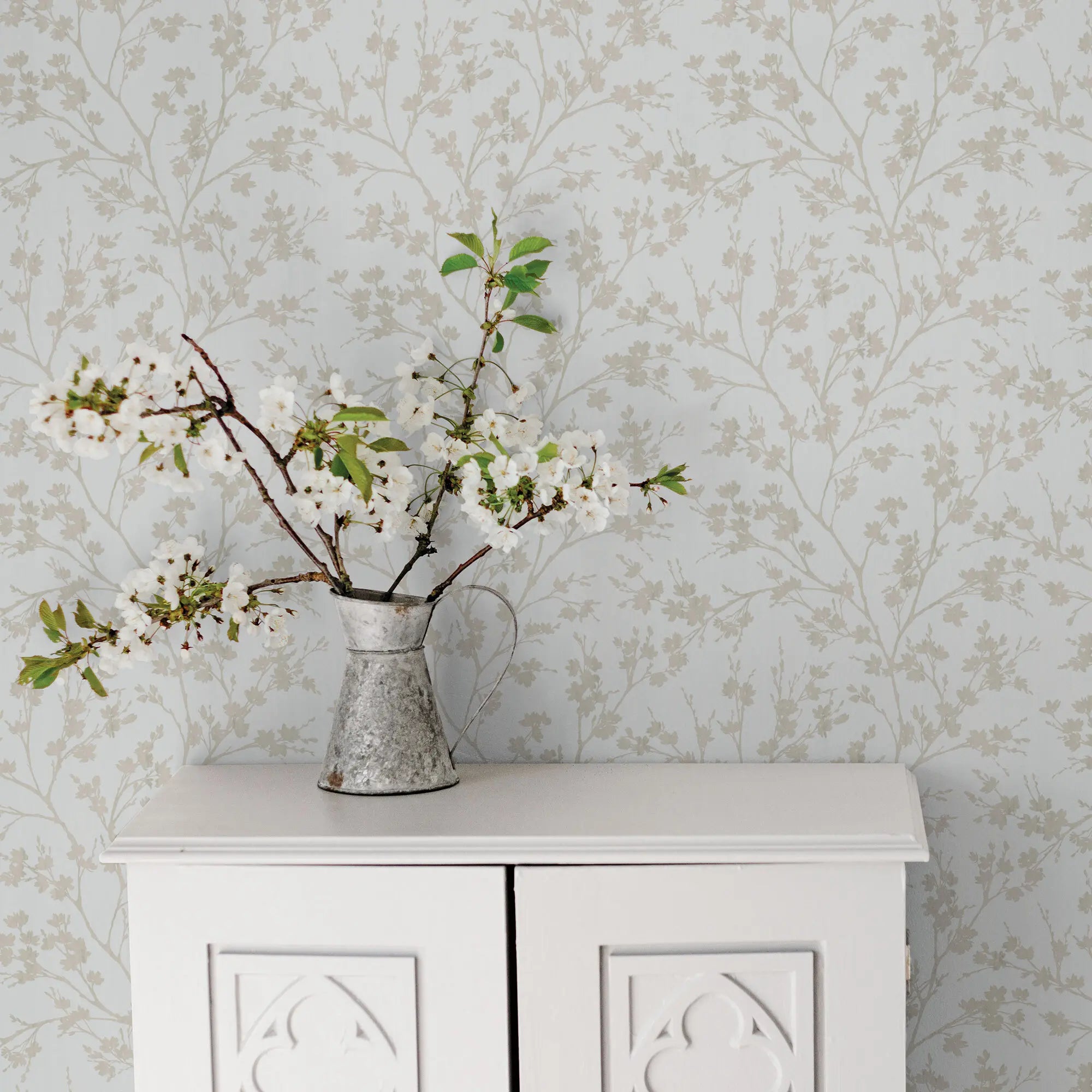 Veiled Branches - Mist fra Nicetapet.dk som indretning. Tapetet danner en elegant baggrund med grene og blomster, bag en hvid skænk med dekorativ vase.